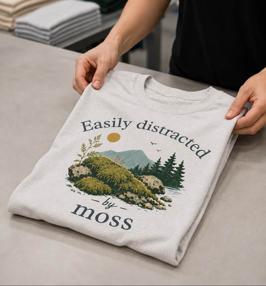 Person holding a folded t-shirt with a nature graphic and text on a table.